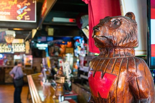 A wooden bear statue with a red "W" stands in a lively bar filled with colorful signs and decorations.
