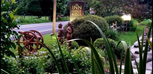 A view of a sign for The Inn at Herr Ridge, surrounded by greenery and decorative statues near a road.