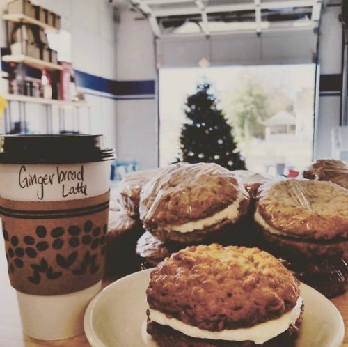 A cozy café scene featuring a gingerbread latte and a plate of cream-filled cookies, with a Christmas tree in the background.