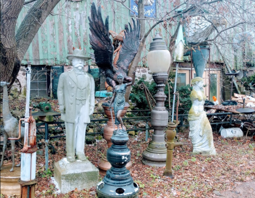 A collection of vintage statues and lamps in a cluttered outdoor space, surrounded by overgrown foliage.