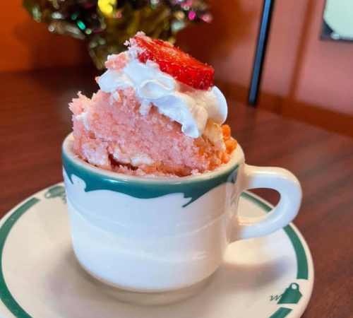 A small cup filled with pink cake topped with whipped cream and a slice of strawberry, resting on a decorative plate.
