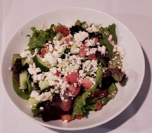 A fresh salad in a white bowl, featuring mixed greens, diced tomatoes, cucumbers, and crumbled feta cheese.