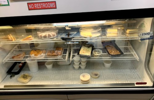 Display case filled with various desserts, including cakes, pastries, and cups of ice cream. Sign reads "No Restrooms."