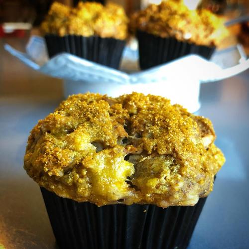 Close-up of a golden-brown muffin with a crumbly topping, set against a blurred background of more muffins.