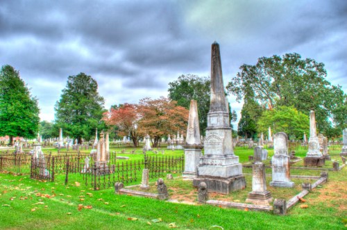 A serene cemetery scene with various gravestones and monuments surrounded by trees under a cloudy sky.