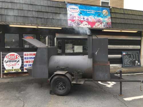 A black smoker grill on a trailer outside a restaurant, with smoke rising and a sign for ice cream in the background.