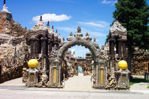 Colorful, ornate archway made of stones and shells, leading into a whimsical, artistic landscape under a blue sky.