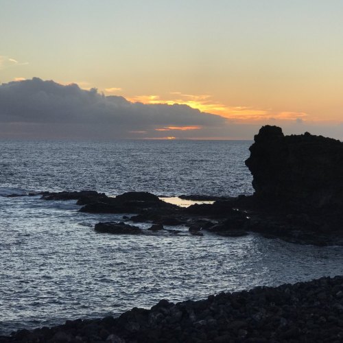 Silhouette of a rocky coastline at sunset, with clouds and a calm sea reflecting warm colors.