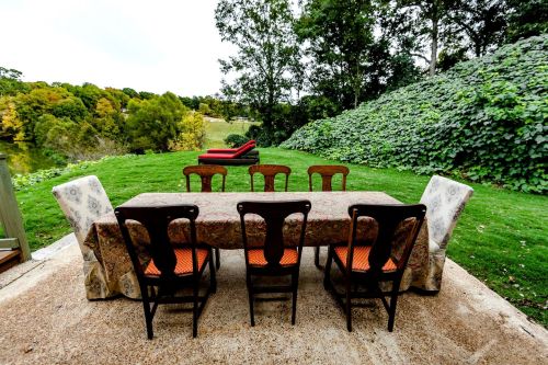 A dining table with chairs set outdoors, overlooking a green landscape and a red lounge chair in the background.
