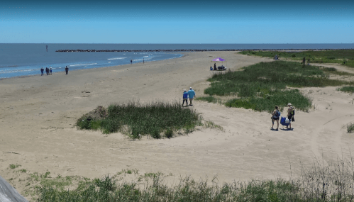 A sandy beach with people walking, grassy areas, and a distant jetty under a clear blue sky.