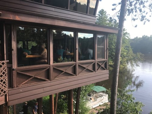 A lakeside building with large windows, surrounded by trees, overlooking a calm water scene with a boat docked nearby.