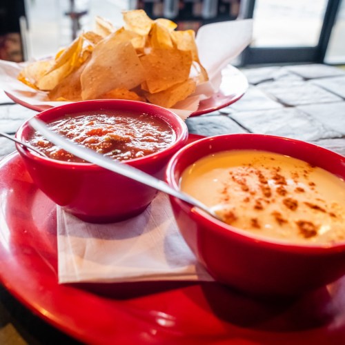 A plate with red bowls of salsa and cheese dip, accompanied by a side of tortilla chips.