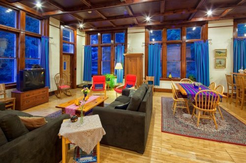 Cozy living room with wooden beams, large windows, red chairs, a dining table, and colorful decor.