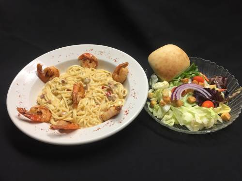 A plate of shrimp pasta with a side salad and a bread roll on a black background.