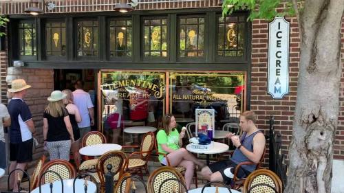 A busy ice cream shop with a line outside, outdoor seating, and a sign reading "Ice Cream" on the wall.