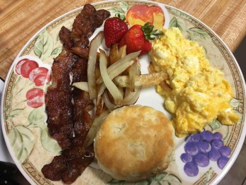 A plate of breakfast featuring bacon, scrambled eggs, fried potatoes, a biscuit, and strawberries.