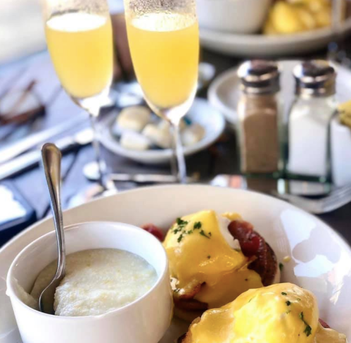 A plate of eggs Benedict with a side of grits, accompanied by two mimosas and condiments on a table.