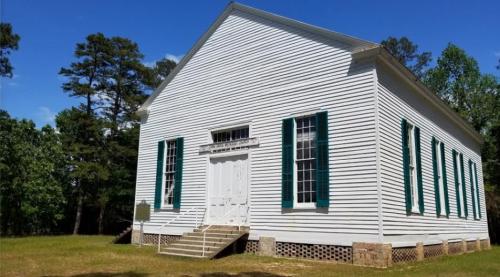 Historic white building with green shutters, surrounded by trees and grass, under a clear blue sky.