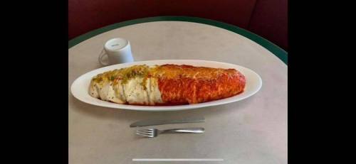 A large, colorful burrito topped with sauce and garnished, served on a white plate with a fork beside it.
