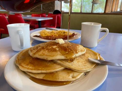 A stack of pancakes drizzled with syrup, served with coffee and a plate of eggs and bacon in a diner setting.