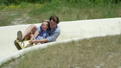A man and a child joyfully ride down a curved slide, surrounded by grass and greenery.