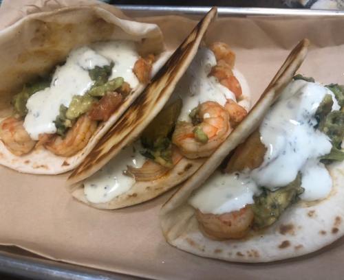 Three shrimp tacos on a tray, topped with guacamole and a creamy sauce, served on soft tortillas.