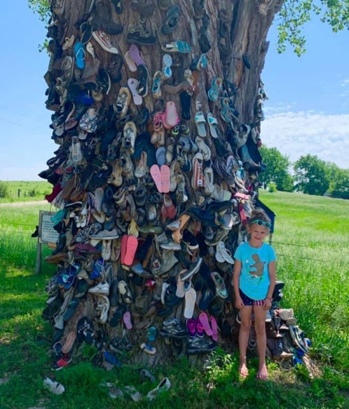 A girl stands beside a tree covered in various shoes, with a green field and blue sky in the background.