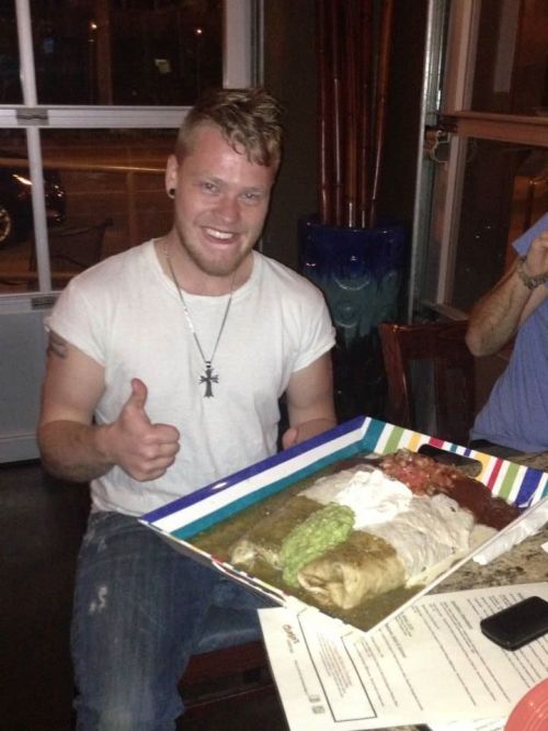 A smiling man gives a thumbs up while holding a large platter of colorful burritos and toppings in a restaurant.