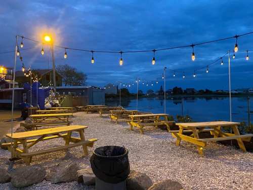 Outdoor seating area with yellow picnic tables, string lights, and a view of a calm water body at dusk.