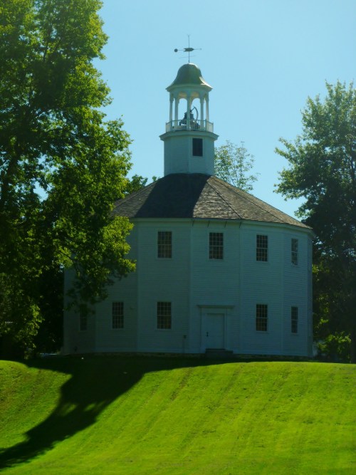 A white, round church with a bell tower, surrounded by green grass and trees under a clear blue sky.