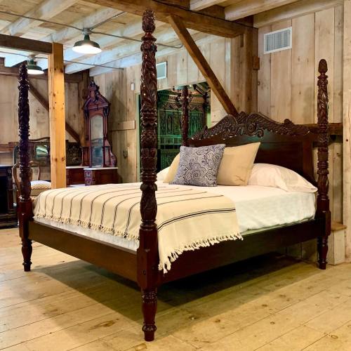 A wooden four-poster bed with intricate carvings, featuring a white blanket and decorative pillows, in a rustic room.