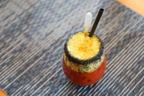 A colorful drink in a glass with a black rim, topped with foam and a straw, placed on a textured surface.