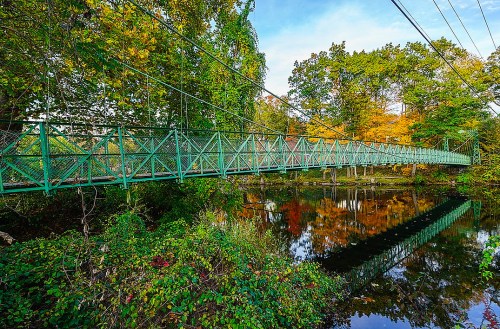 A green suspension bridge spans a calm river, surrounded by trees with autumn foliage reflecting in the water.