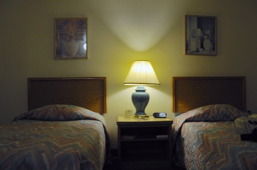 A dimly lit hotel room with two twin beds, a lamp, and framed pictures on the wall.