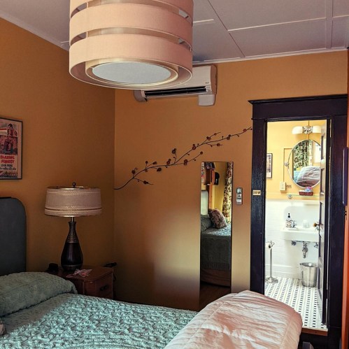 Cozy bedroom with yellow walls, a lamp, and a mirror reflecting a bathroom with vintage decor.