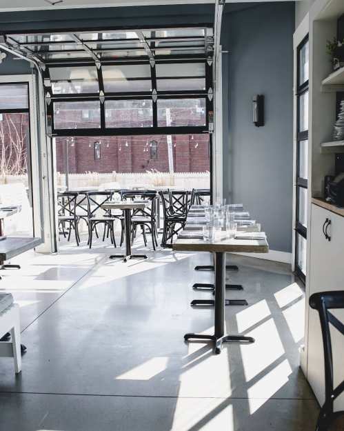 A bright, modern restaurant interior with tables and chairs, featuring large garage doors and concrete flooring.