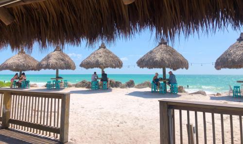Beach scene with thatched umbrellas and turquoise tables, people enjoying the ocean view on a sunny day.