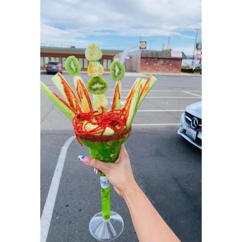 A hand holds a colorful fruit and vegetable bouquet in a glass, featuring celery, kiwi, cucumber, and chili powder.