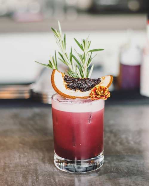 A vibrant cocktail garnished with rosemary, dried orange, and a flower, served in a clear glass on a bar counter.