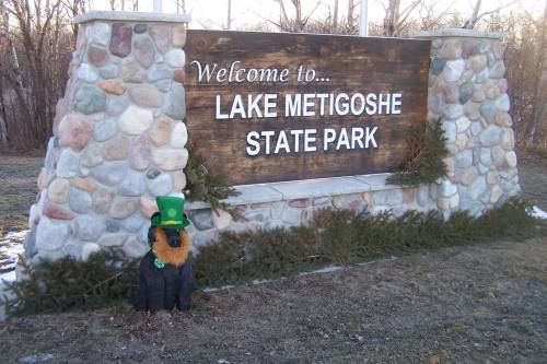 Welcome sign for Lake Metigoshe State Park, featuring a stone structure and a dog statue wearing a green hat.