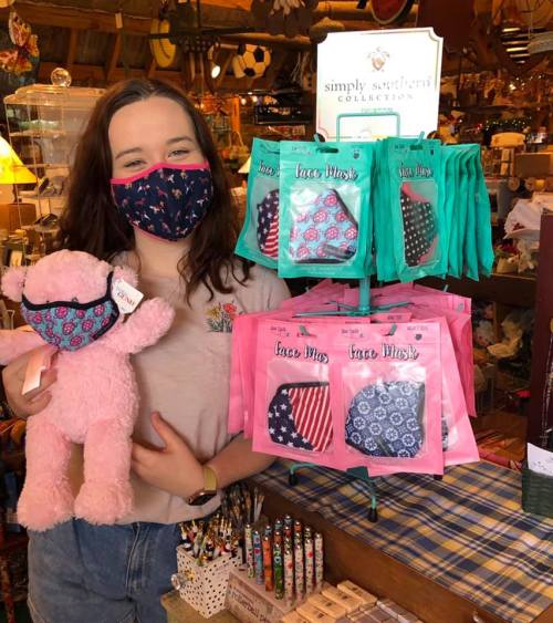 A young woman wearing a mask holds a pink stuffed animal, standing in front of a display of colorful face masks.