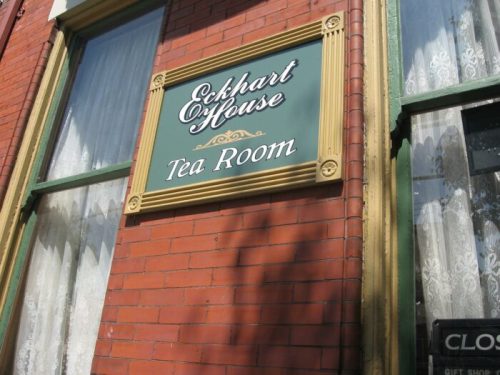 Sign for "Eckhart House Tea Room" on a brick wall, featuring decorative gold accents and lace curtains in the background.