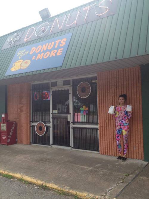 A woman in a colorful donut-patterned outfit stands outside a donut shop with an "OPEN" sign.