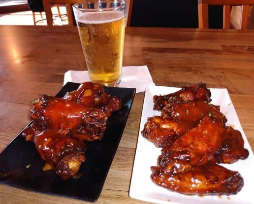 Two plates of glazed chicken wings with a pint of beer on a wooden table.