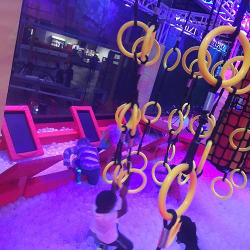 Children play on colorful gym equipment surrounded by soft balls in a vibrant, illuminated indoor play area.