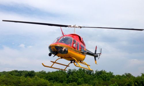 A red and yellow helicopter flying above green trees under a cloudy sky.