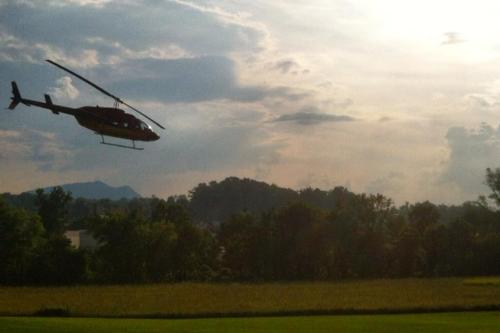 A helicopter flies over a green field with trees and mountains in the background under a cloudy sky.