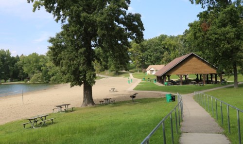 A scenic park with a sandy beach, picnic tables, and a pavilion near a calm lake surrounded by trees.