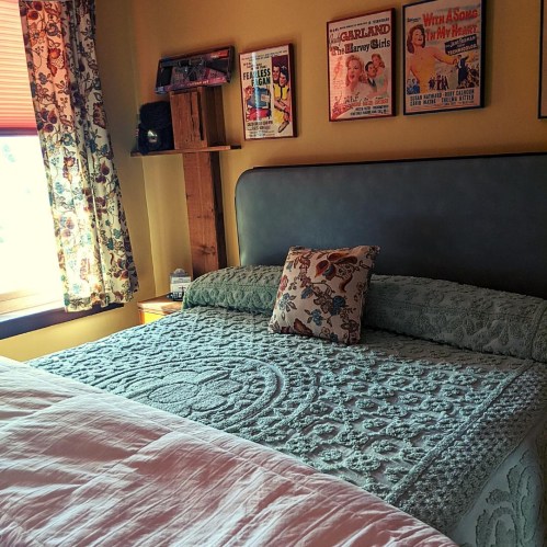 Cozy bedroom with a green quilted bedspread, decorative pillow, and vintage movie posters on the wall. Natural light from the window.