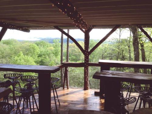 Cozy wooden deck with bar stools, overlooking lush green trees and distant mountains under soft lighting.